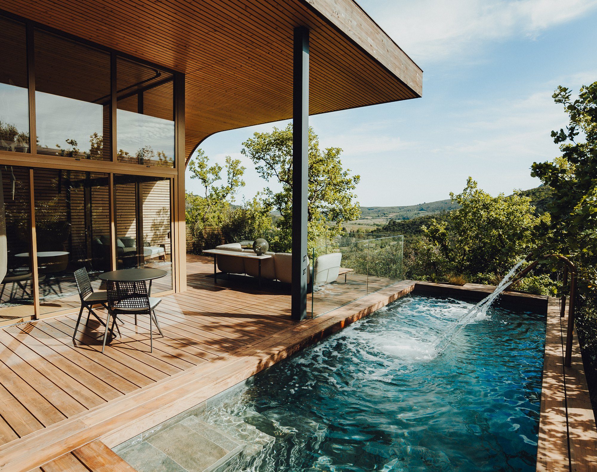La piscine privée d'un lodge Souki vue dans sa longueur depuis la terrasse en bois, un canon de massage en action et les collines au fond