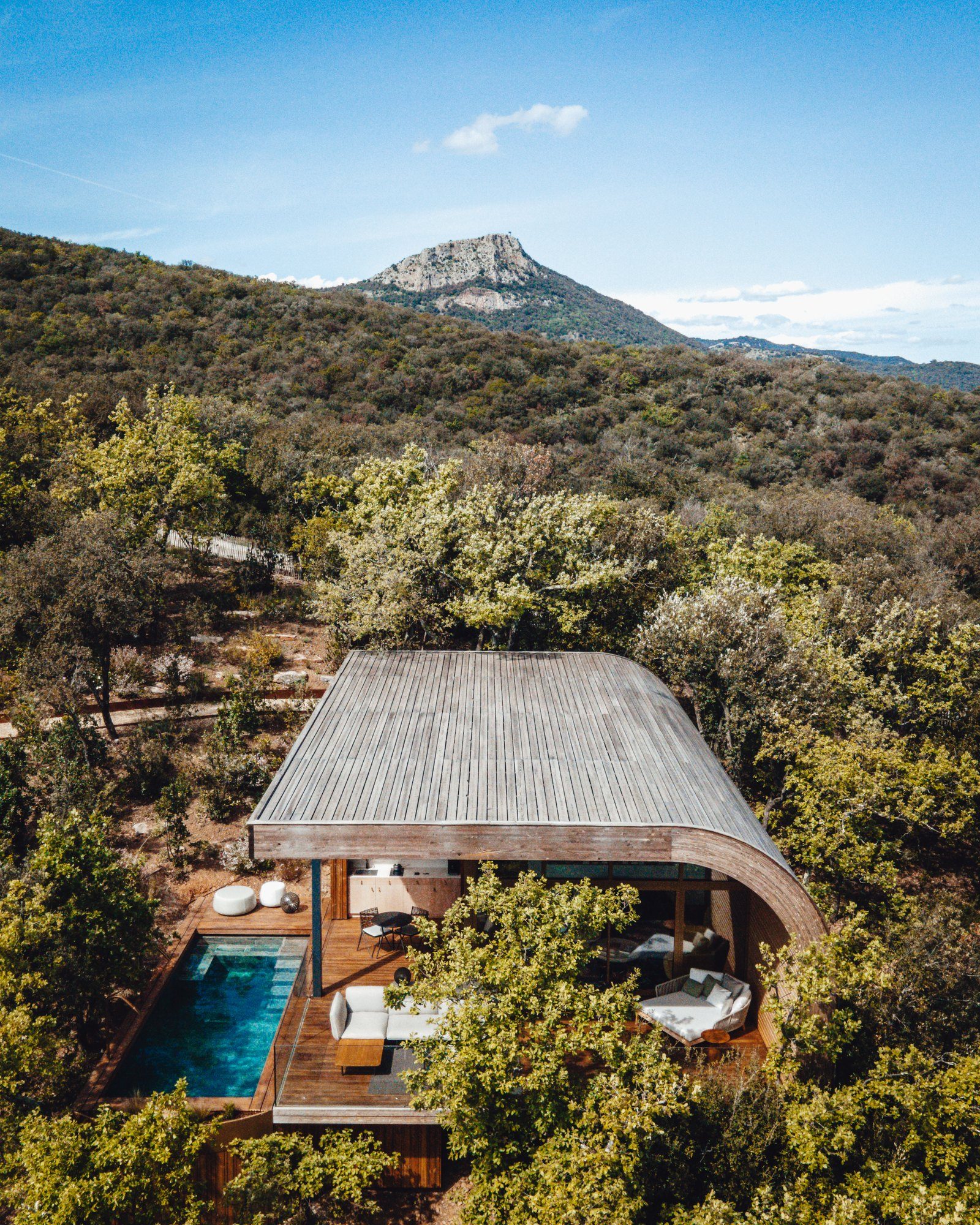 Un lodge Souki vu du ciel à Cabrières : sa toiture courbe en bois grisé posée dans la chênaie, sa piscine privée le long de la terrasse, et le Pic de Vissou au fond