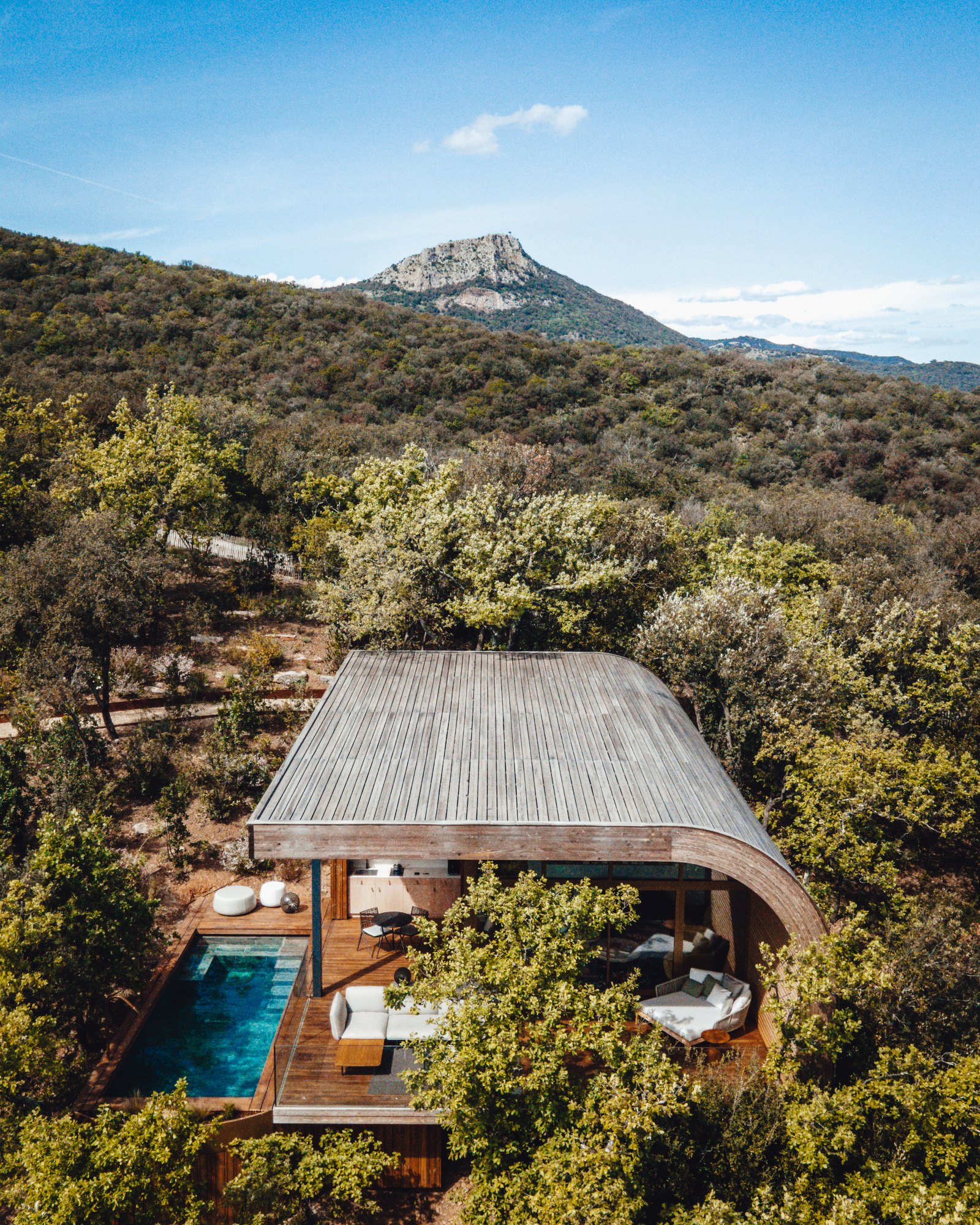 Un lodge Souki vu du ciel à Cabrières : sa toiture courbe en bois grisé posée dans la chênaie, sa piscine privée le long de la terrasse, et le Pic de Vissou au fond