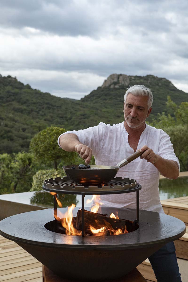 Gilles cuisine au brasero sur la terrasse, flammes vives, collines de Cabrières en arrière-plan