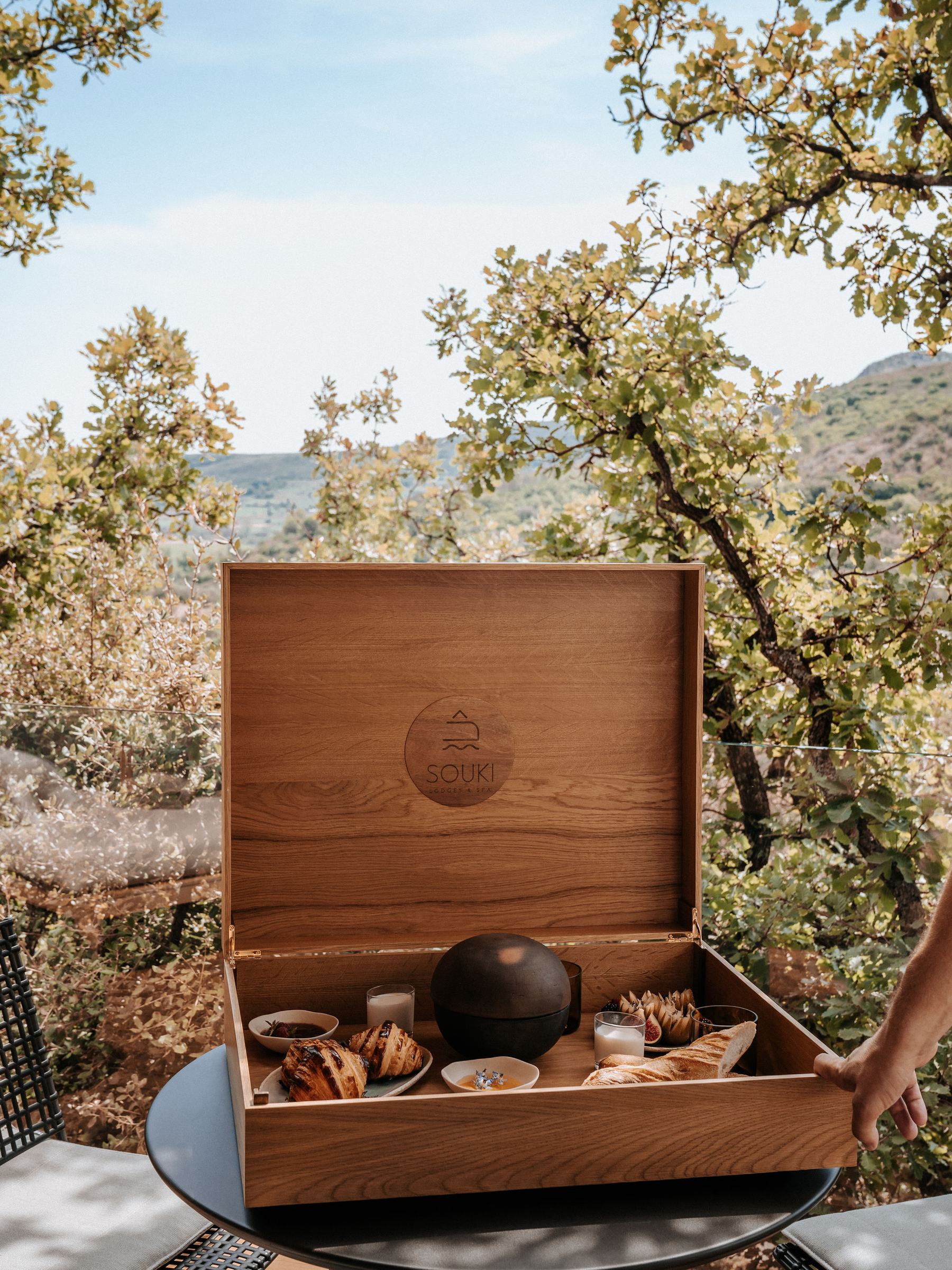 La grande malle en bois ouverte, son couvercle gravé Souki : viennoiseries, pain, confitures, figues et yaourts, dehors sous les chênes